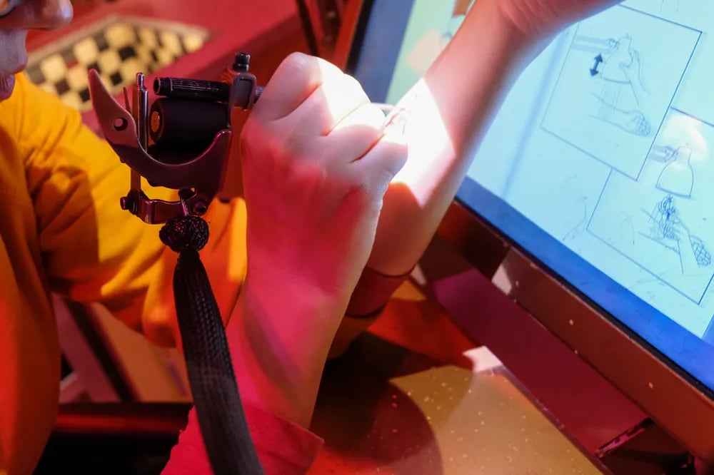 Tattoo machine in a gloved hand beside a drawing tablet showing the design